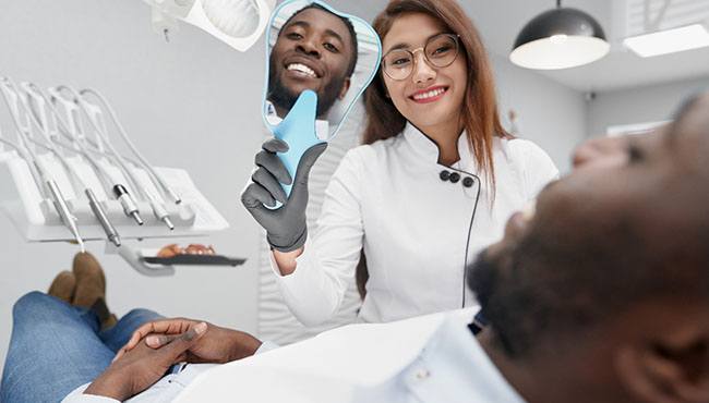 A dentist holding up a handheld mirror to show a male patient his new smile with Lumineers