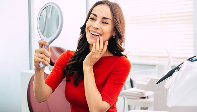 A woman wearing a red blouse and looking at her smile in the mirror while at the dentist’s office