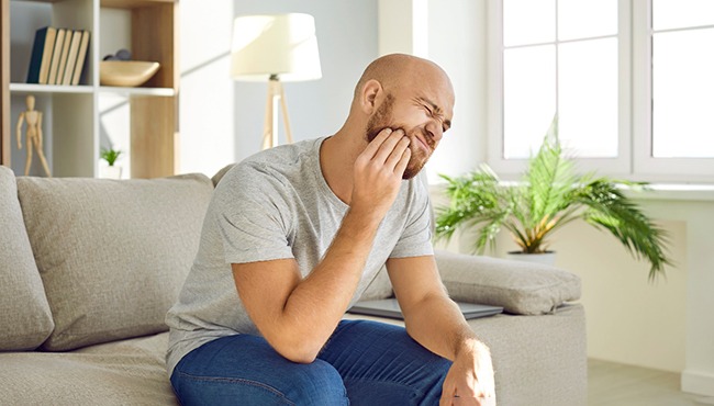 Man in grey shirt sitting on couch with tooth pain