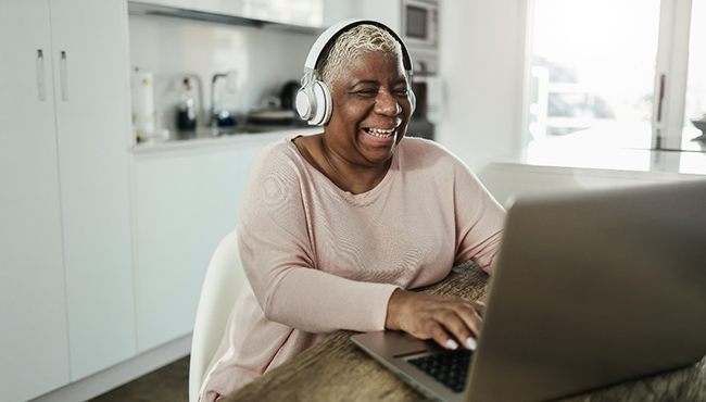 a smiling person sitting at a table with headphones on using their laptop